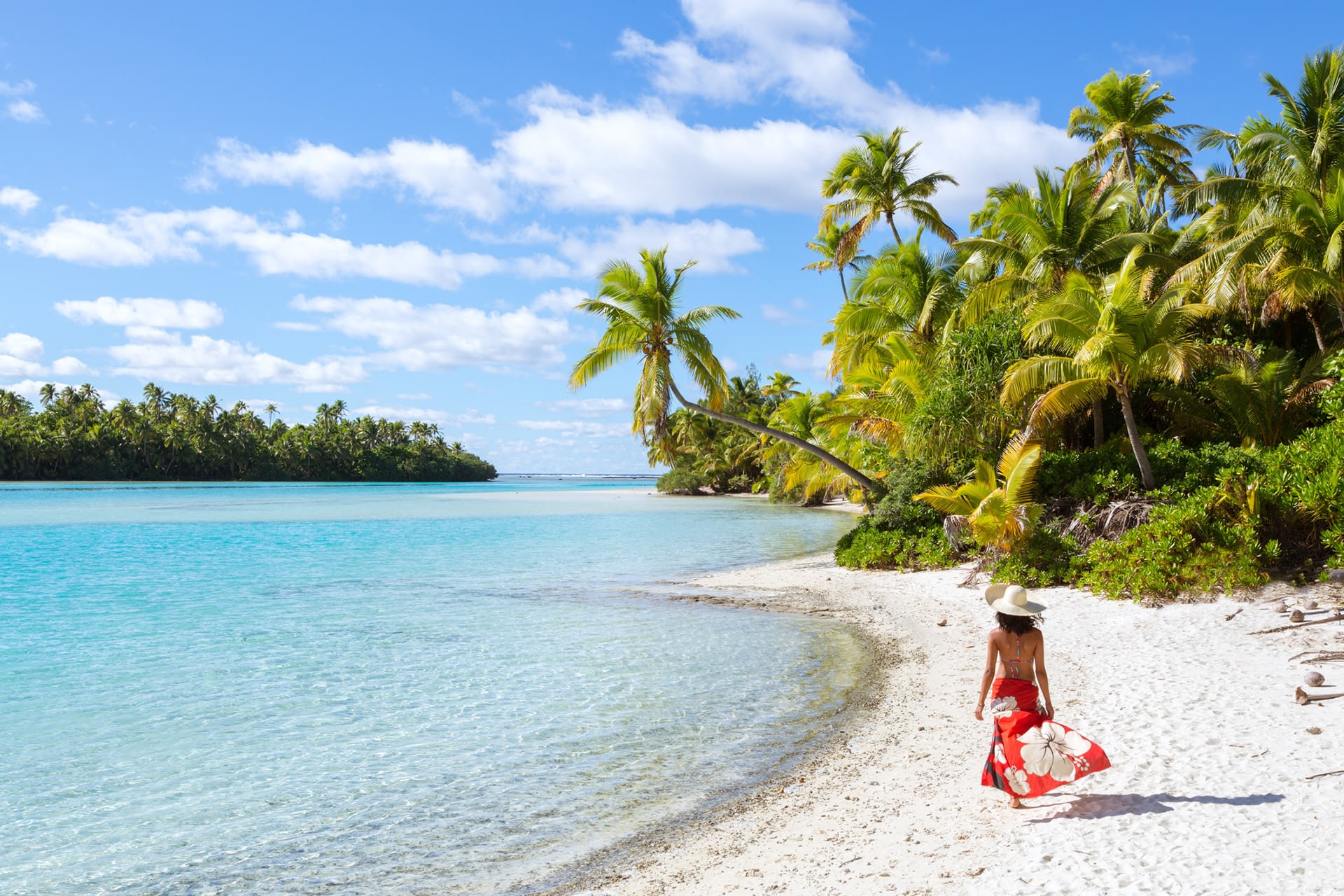 Woman in bikini with sarong and straw hat walking on the famous beach of Tapuaetai, a small islet in the south-east of the lagoon of Aitutaki, Cook islands.